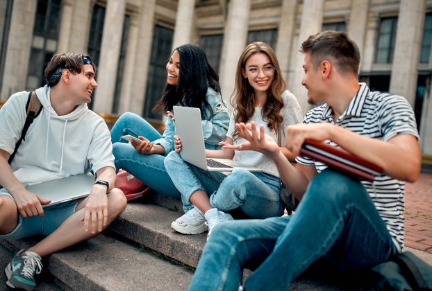 A group of students with laptops sit on the steps near the campus and communicate.