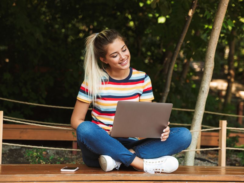 Portrait of an excited young teenage girl sitting on a bench at the park, using laptop computer on her lap