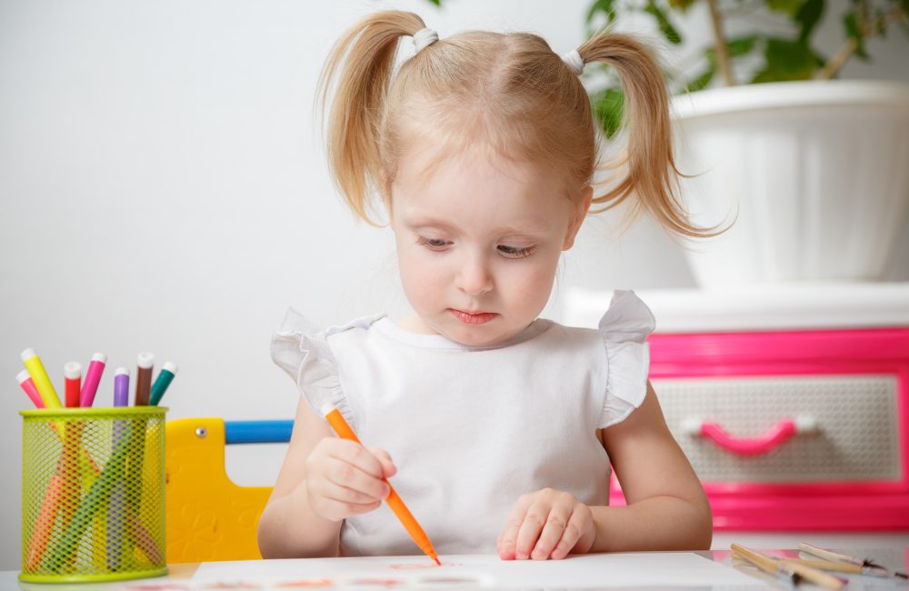 little beautiful girl 3-4 years old with two ponytails in a white T-shirt paints at the table and smiles. quarantined children's creativity due to coronavirus